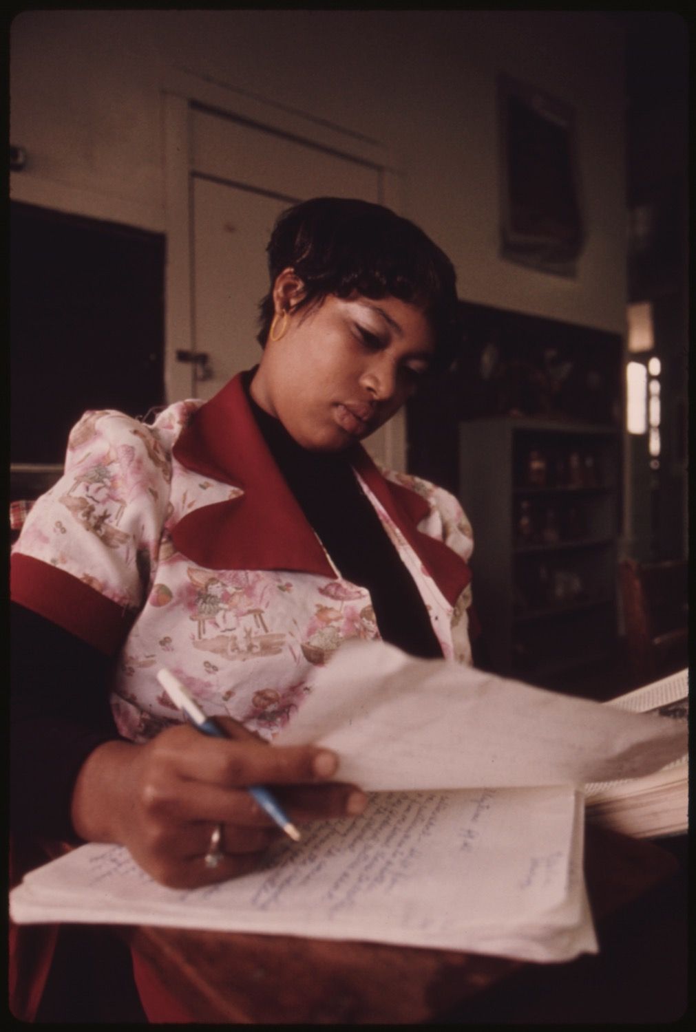 A woman is writing on a sheet of paper with a blue pen, seated at a table in a dimly lit room.