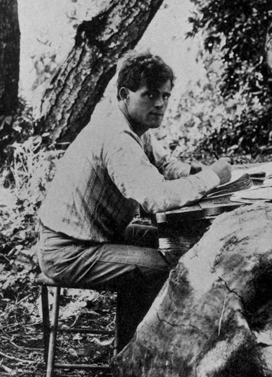 A black and white photograph of a man seated outdoors, writing or drawing on a flat surface.