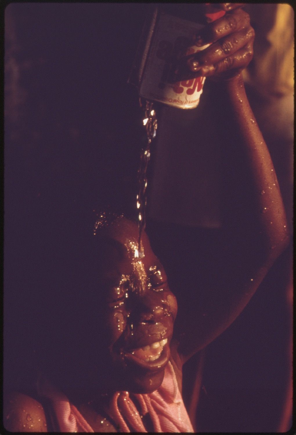 Close-up of a person with dark skin having a dark liquid poured over their head, with droplets glistening in the light.