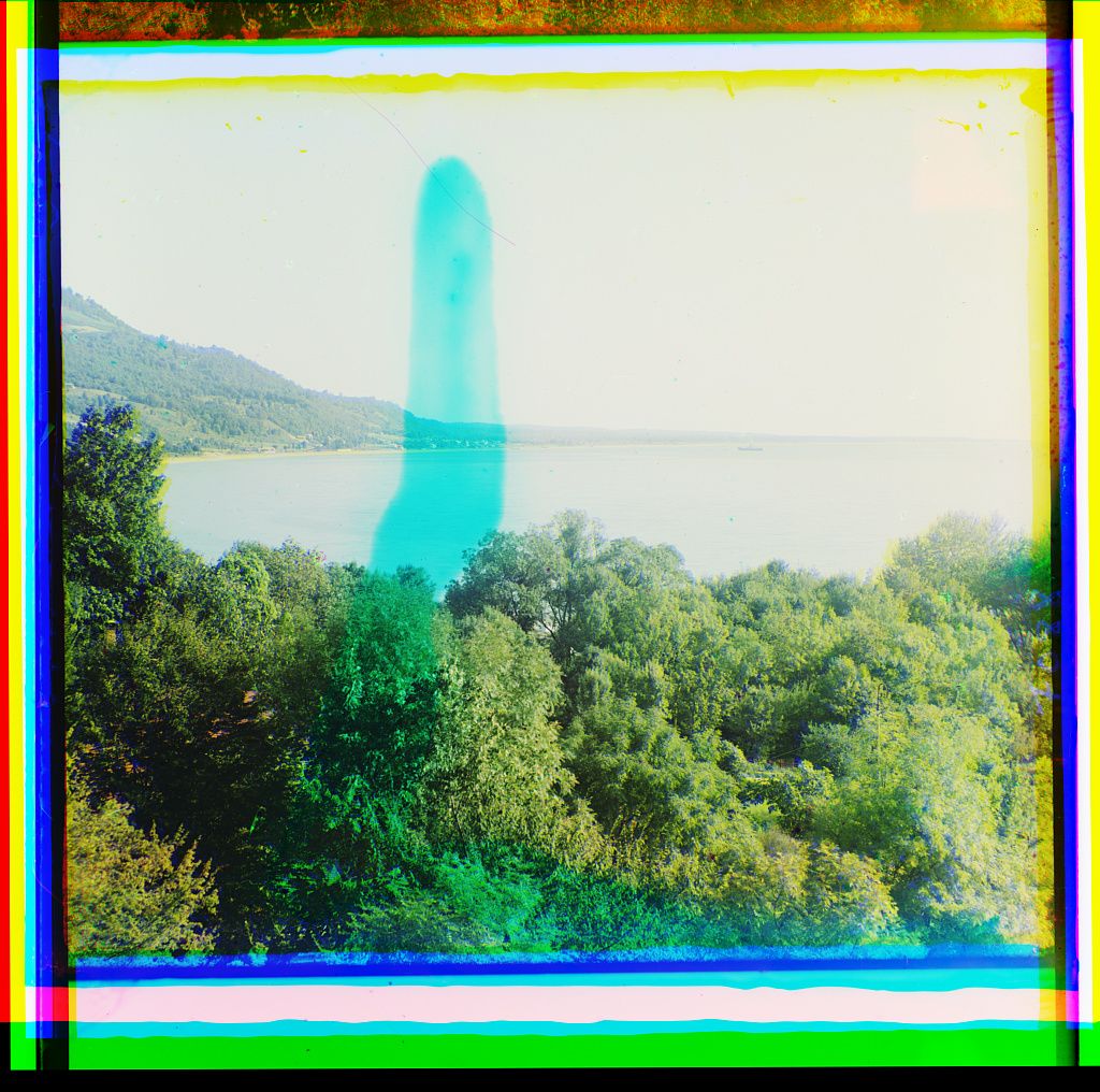 A vibrant, slightly distorted view of a water fountain shooting high into the air over a lake, with trees in the foreground and mountains in the background.