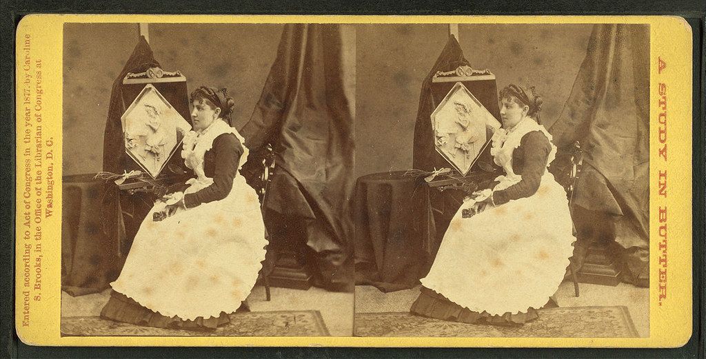Stereoscopic photograph of a woman in a long dress holding a fan, seated in a chair with dark curtains in the background.