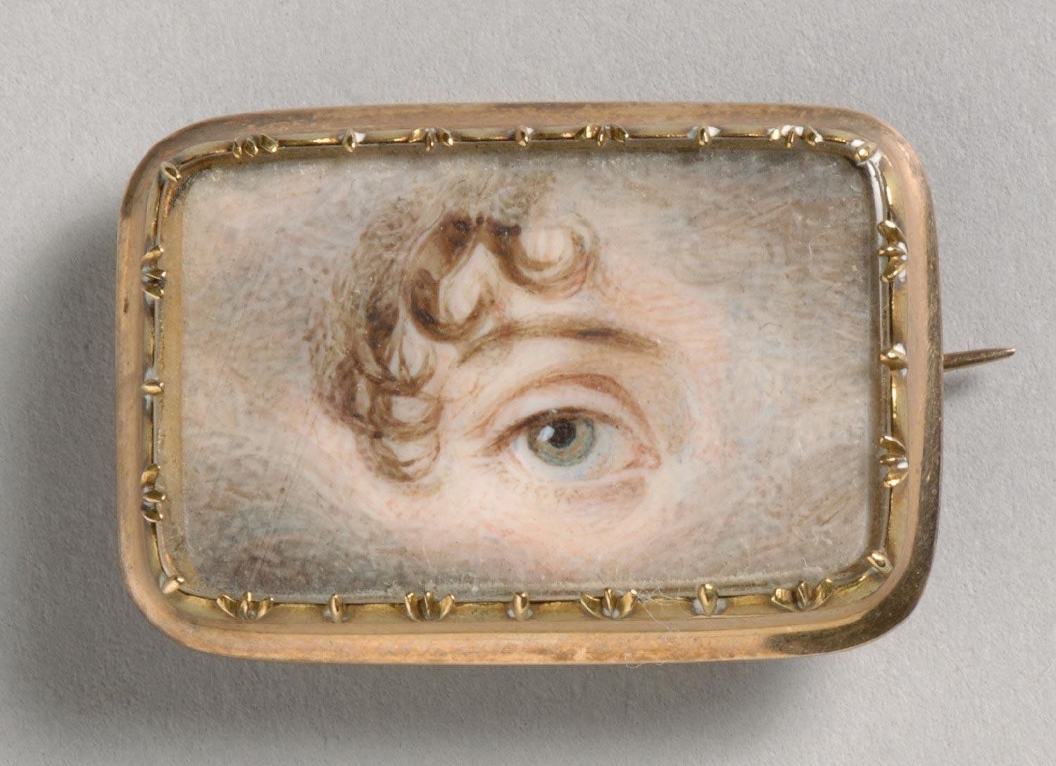 Close-up of a rectangular gold brooch with a miniature painting of a blue eye and curly brown hair.