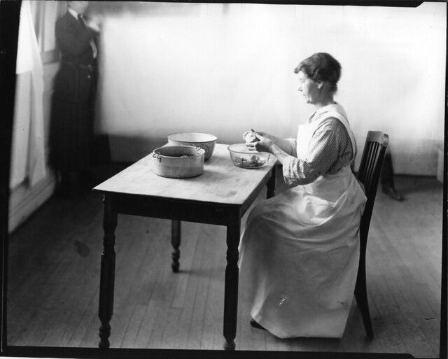 A black and white photograph of a woman seated at a table, working with bowls in a simple room.