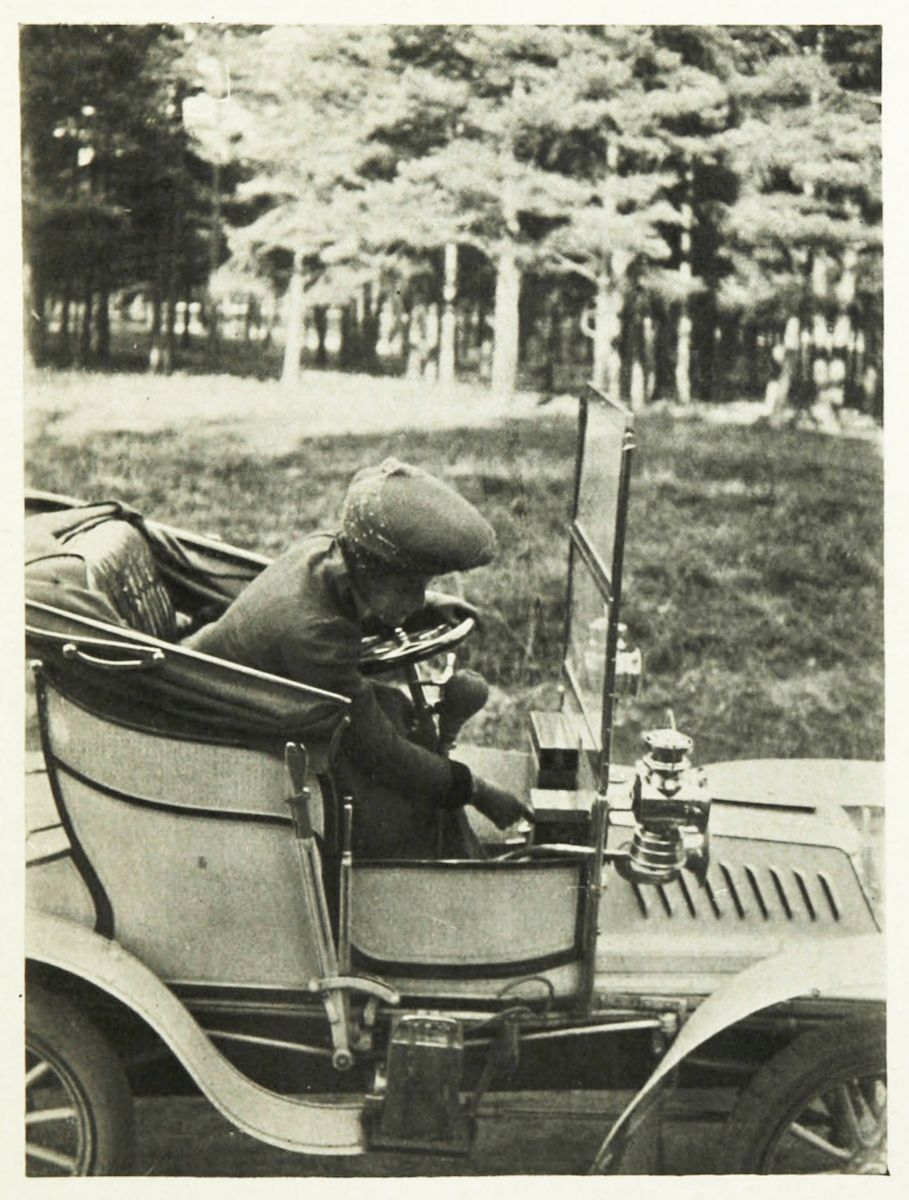A vintage black and white photograph of a person driving an early model car in a grassy area with trees in the background.