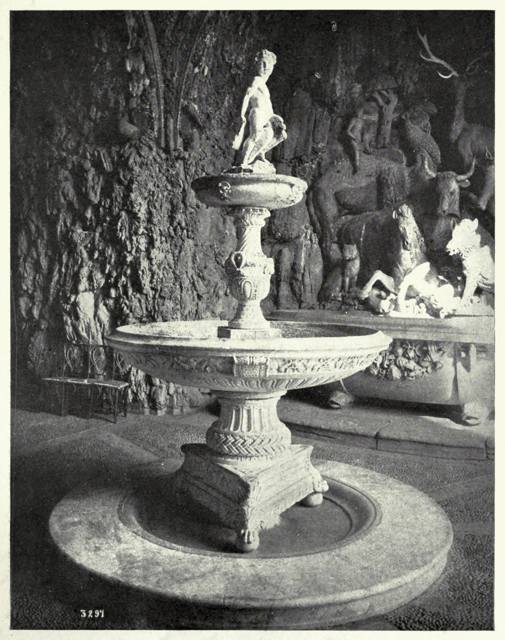 A black and white photograph of a classical-style fountain with a statue on top, set in a garden or historical setting.