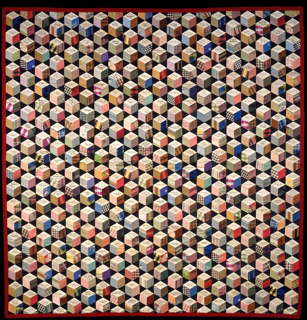 A vintage quilt made of hundreds of hexagonal fabric patches, each with handwritten signatures, arranged in a repeating pattern and bordered by a dark band.