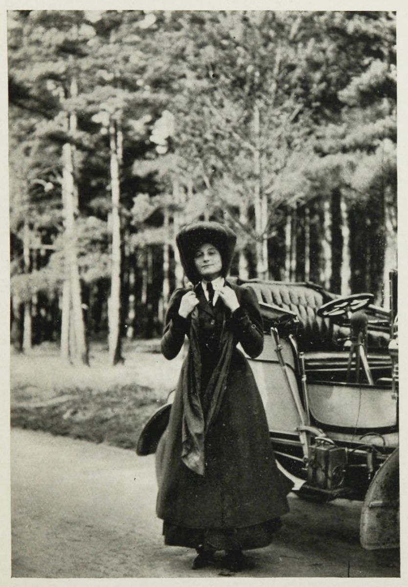 A vintage black and white photograph of a woman working on the front tire of a vintage car in a forest.