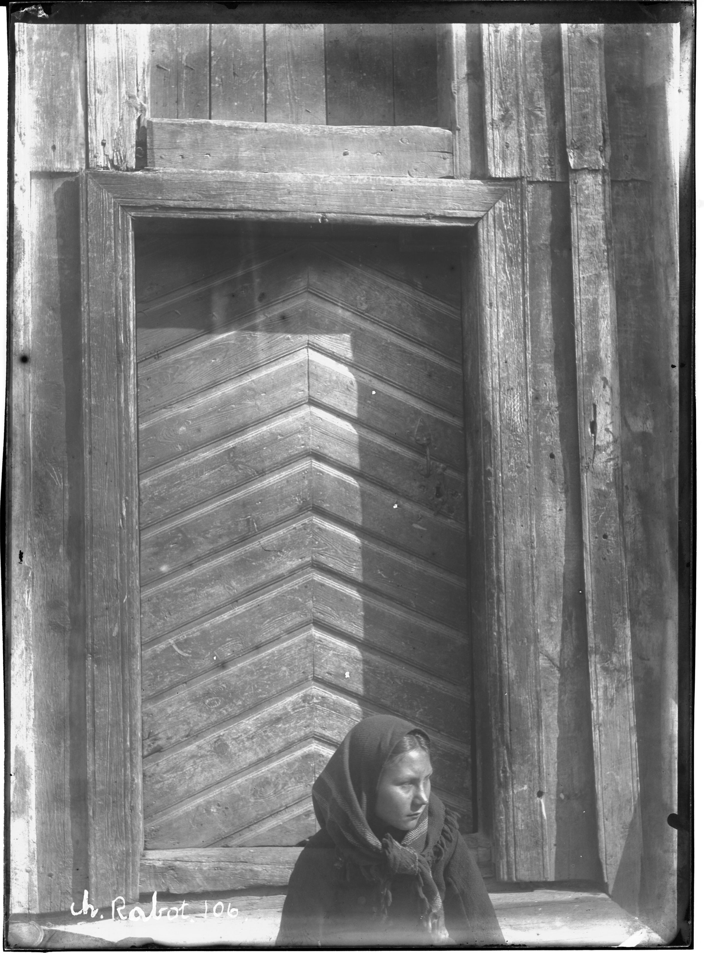 A black and white photograph of a woman in a headscarf standing in front of a wooden door with a diamond pattern.
