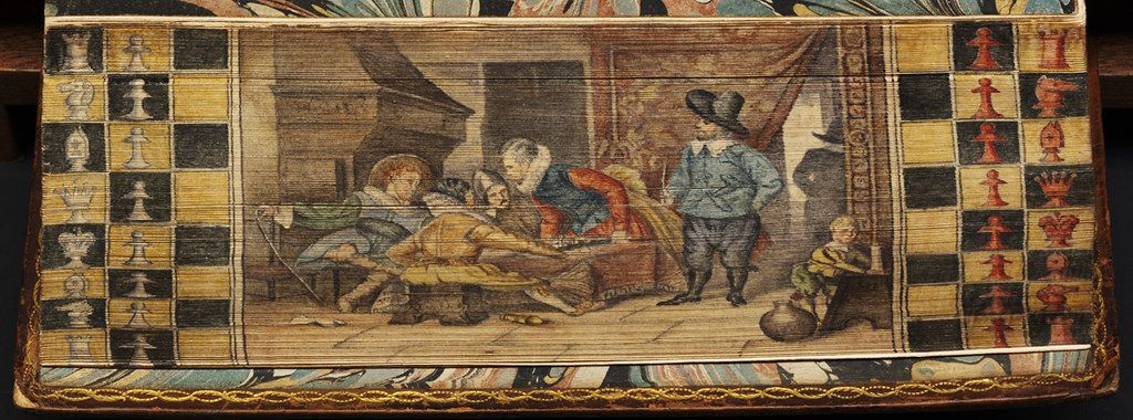A rectangular painting depicting an interior scene with people gathered around a table playing a board game. The painting is framed by a decorative border featuring a checkerboard pattern with chess pieces and a gold trim.