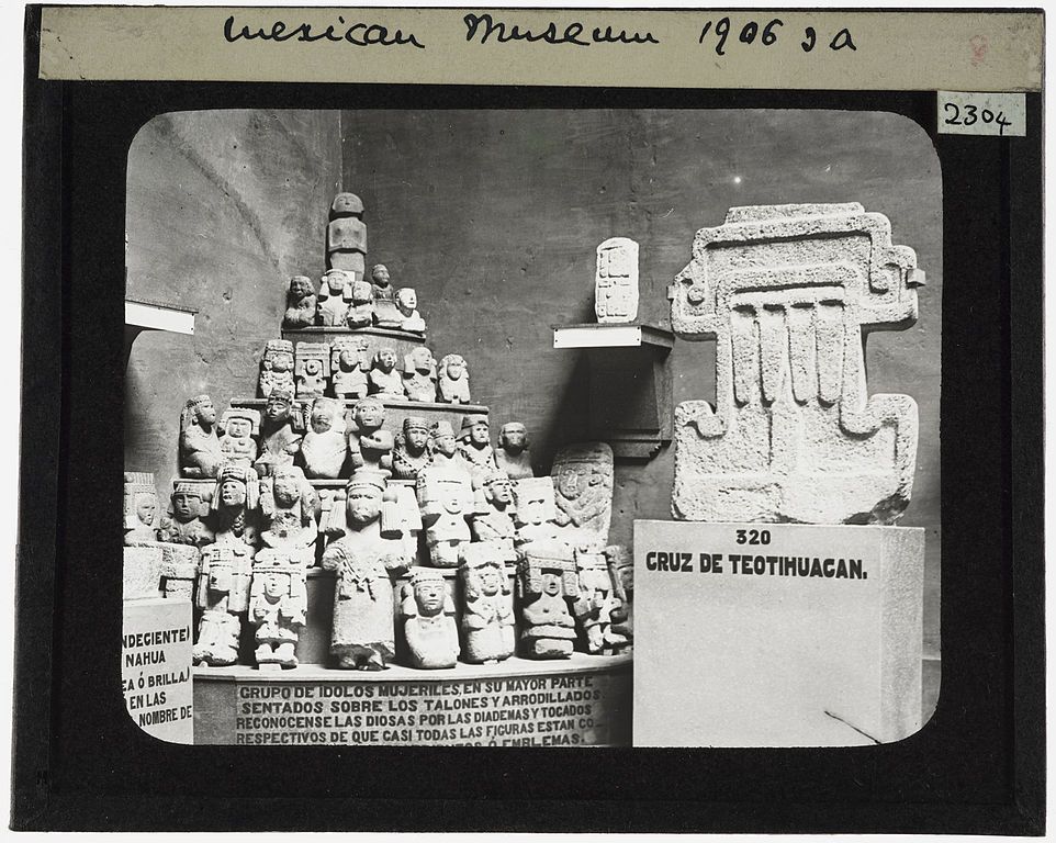 A black and white photograph of a museum exhibit featuring a stone cross adorned with numerous small figures, alongside a collection of similar statues. The exhibit is labeled 'CRUZ DE TEOTIHUACAN' and dated 1906.