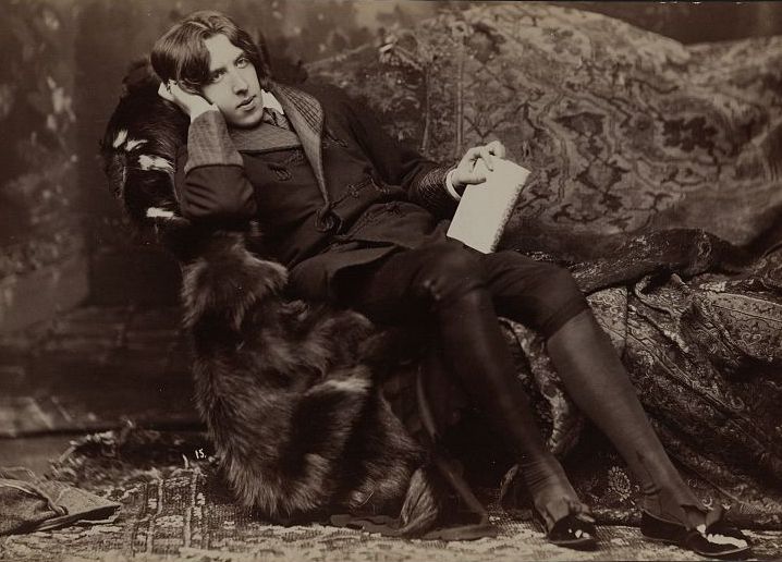 A sepia-toned photograph of a young man reclining in a luxurious chair, holding a book, and looking thoughtful.