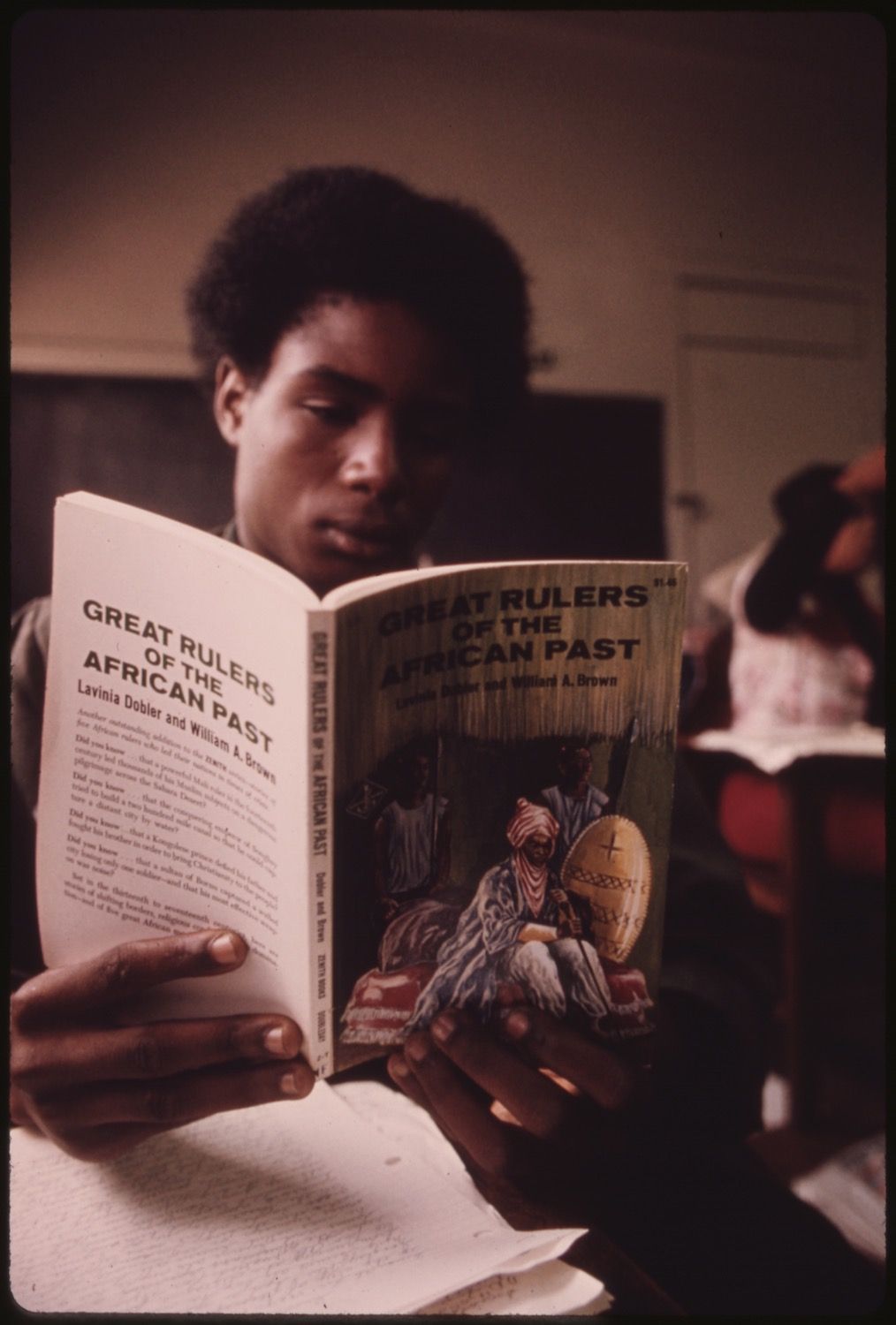 A young Black boy reading a book titled "Great Rulers of the African Past" in a classroom setting.