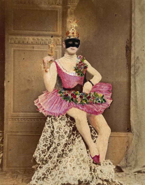 A vintage hand-colored photograph of a person in a theatrical costume, wearing a pink tutu, black mask, and ornate headpiece, seated against a textured wall.