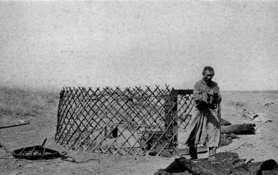 A black and white photograph of a man standing next to a woven fence-like structure in a barren landscape.