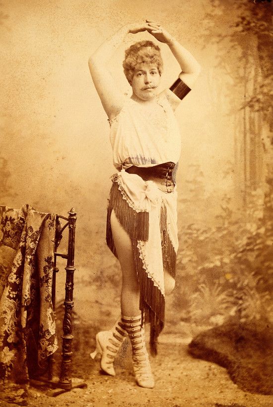 Vintage sepia-toned photograph of a woman in a theatrical costume, posing with one arm raised and wearing a ruffled top, short skirt, stockings, and shoes.