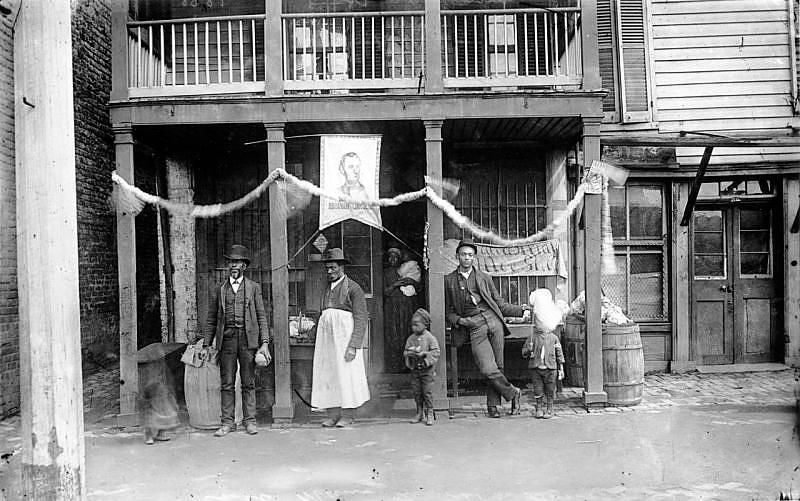 A vintage black and white photograph of a street scene with a row of buildings and a group of people standing in front of one of them.