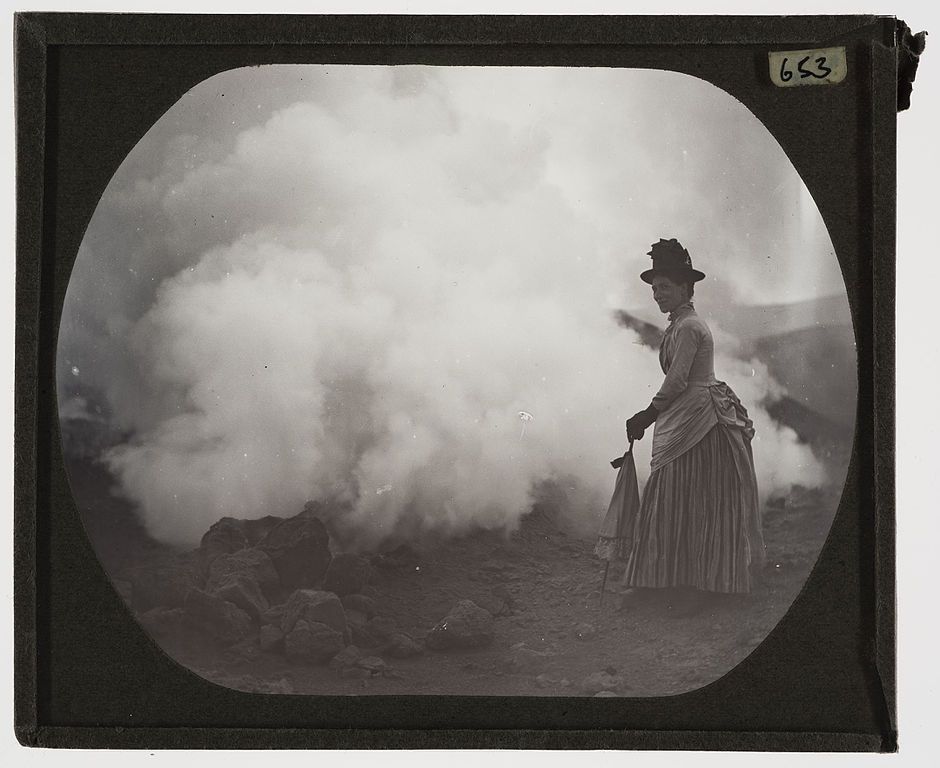 A vintage black and white photograph of a woman in Victorian dress standing in a hazy landscape.