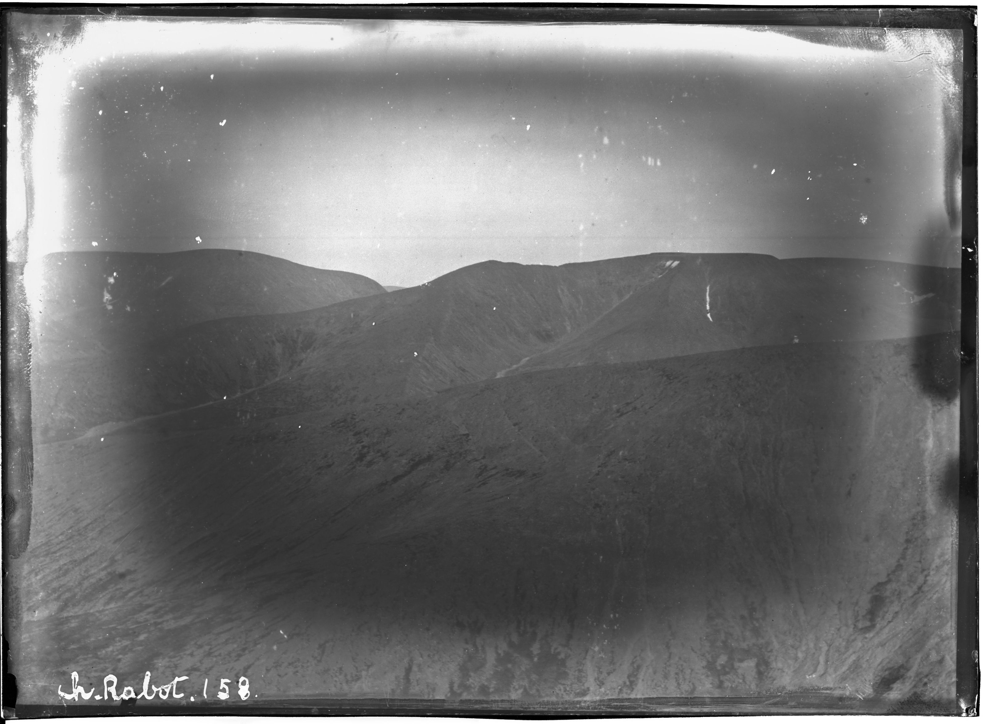 A vintage black and white photograph of a mountain landscape with a bright sky and a dark foreground.