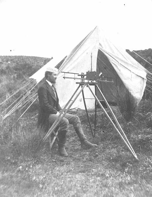 A vintage photograph of a man operating a large camera on a tripod in front of a tent in a grassy field.