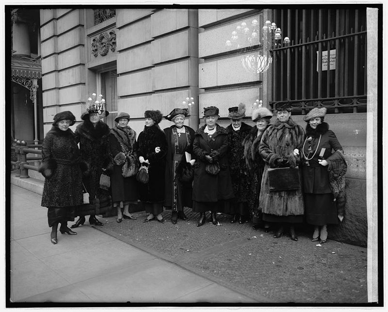 A black and white photograph of ten women standing on a street in front of a building. They are all dressed in long, dark coats and hats, typical of the early 20th century.
