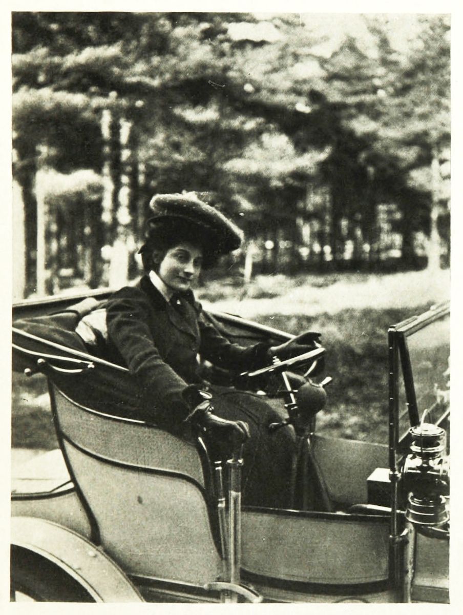 A vintage black and white photograph of a woman driving an early automobile.