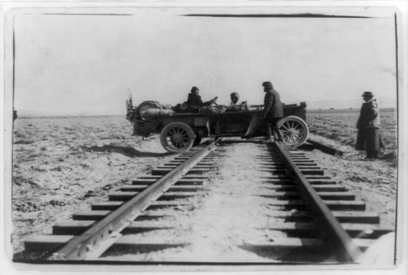 A vintage automobile is stuck on a set of railroad tracks in a vast, open field, with several people gathered around it.