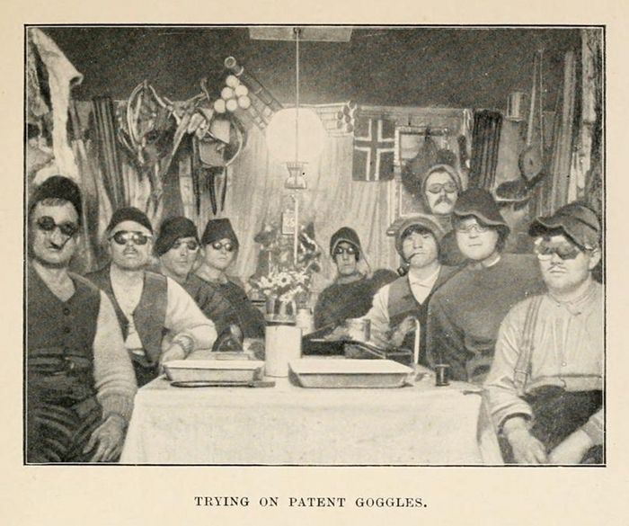 A vintage black and white photograph titled "Trying on Patent Goggles" shows a group of people seated around a table, all wearing large goggles.