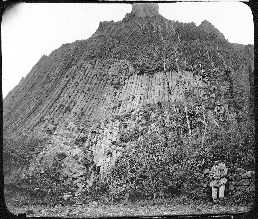 A black and white photograph of a massive cliff face composed of perfectly aligned basalt columns, with a person standing near a stone wall at the base.