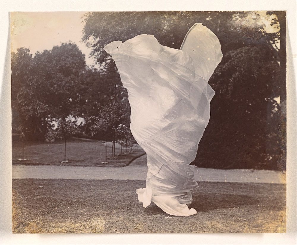 A vintage black and white photograph of a person draped in flowing fabric in a park setting.