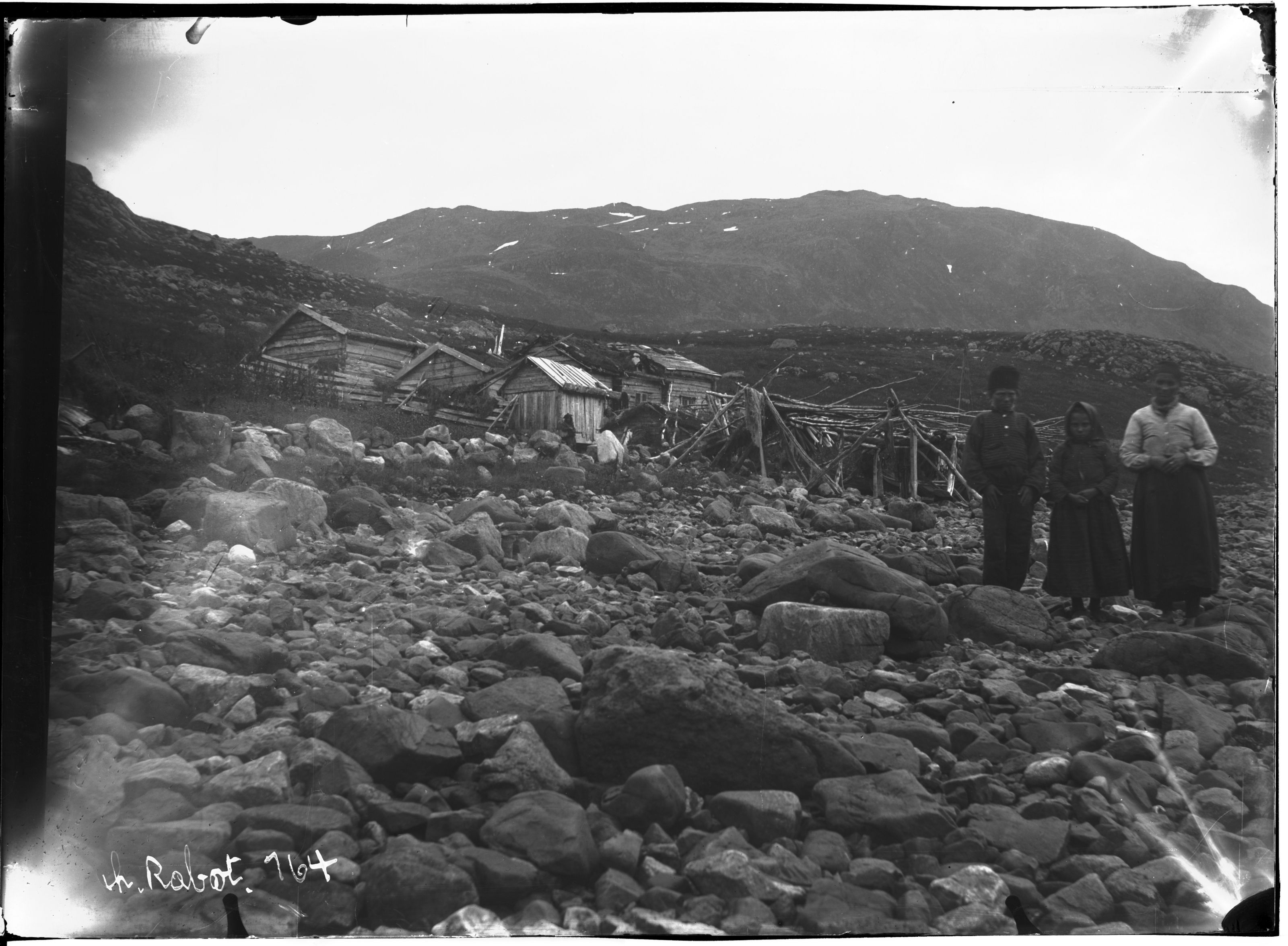 A black and white photograph of a dilapidated building in a rocky landscape with two people standing nearby and a mountain in the background.