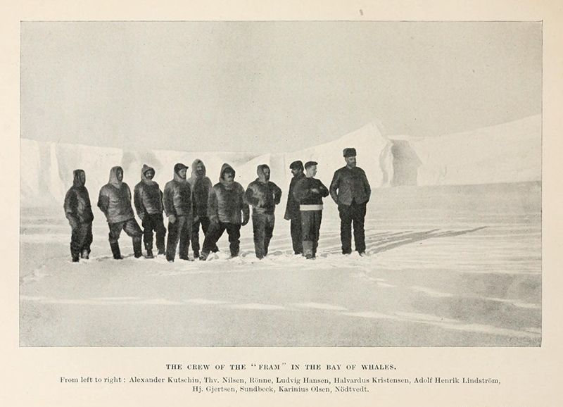 A vintage black and white photograph of a group of men dressed in heavy winter clothing standing in a snowy, arctic landscape. The caption below the image lists the names of the crew members from left to right.