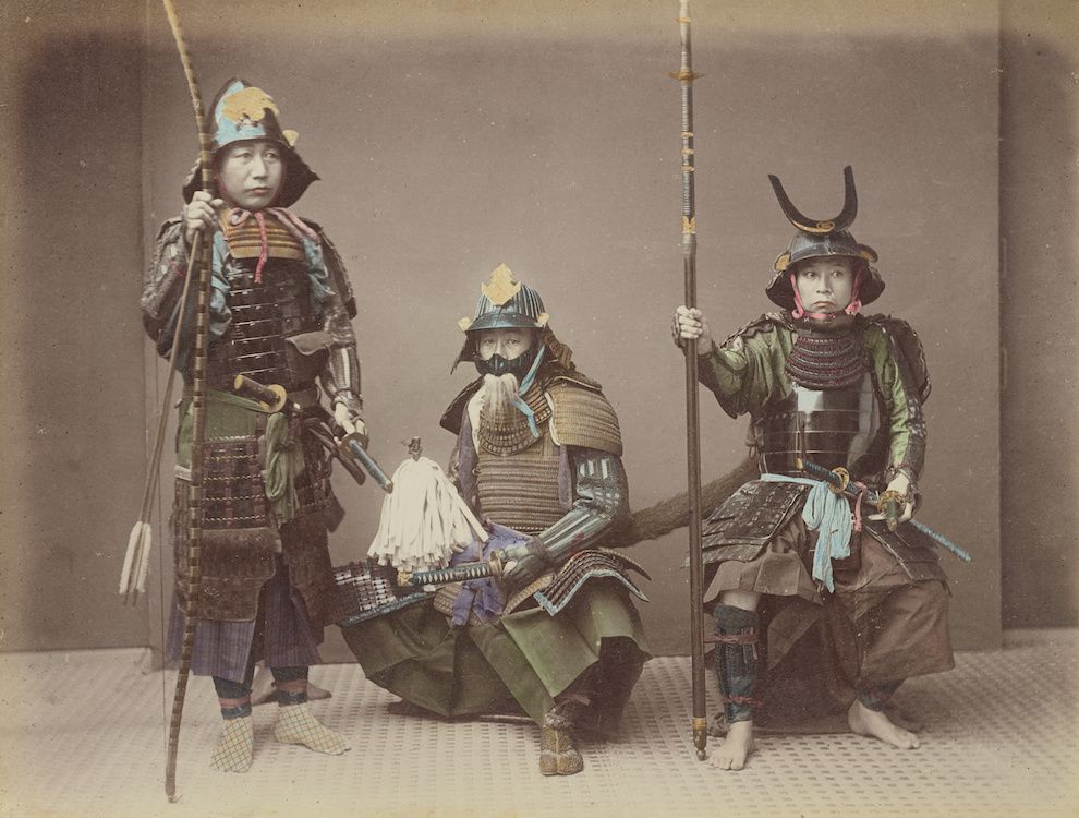 A sepia-toned studio portrait of three samurai warriors in full traditional armor, each carrying a weapon and posed against a plain background.