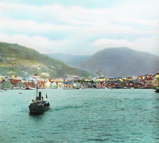 Vintage colorized photograph of a coastal town with a steam boat in the foreground and hills in the background.