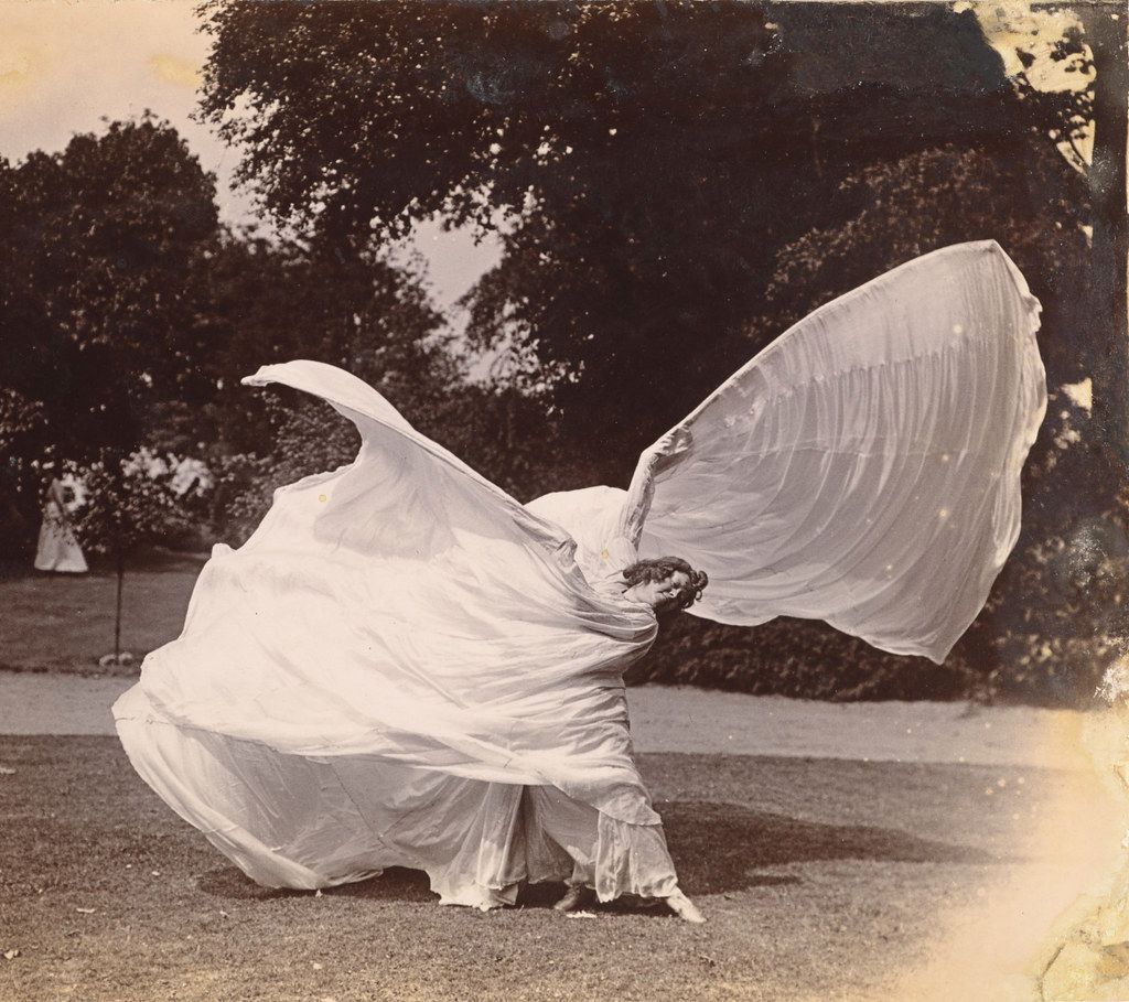 A vintage black and white photograph of a person enveloped in a large, flowing white fabric, with trees and grass in the background.