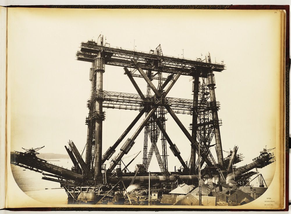 A vintage black and white photograph of a massive industrial structure under construction, featuring a complex network of metal beams and supports over water.