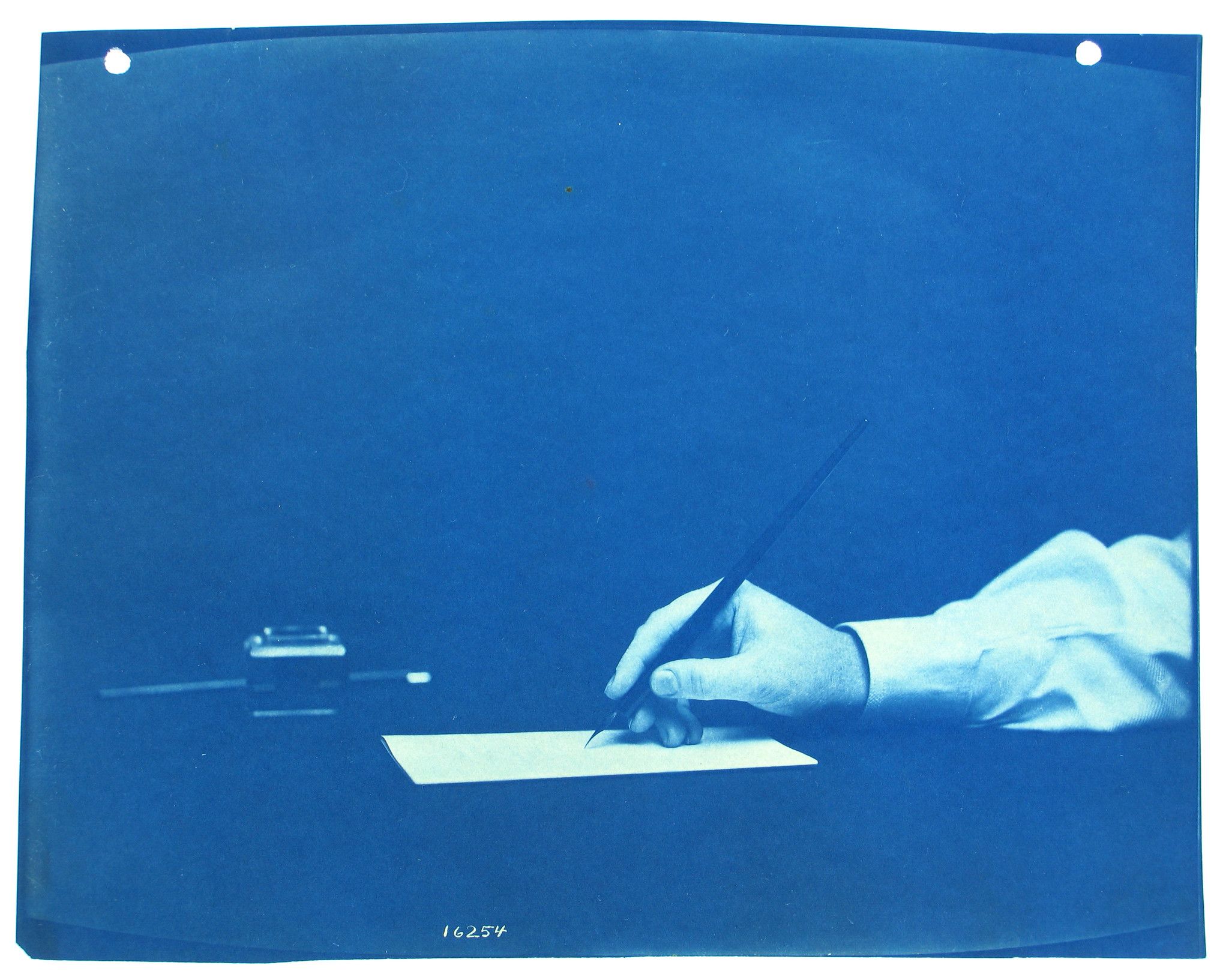 Cyanotype photograph of a hand writing on a piece of paper with an inkwell and pen holder in the background.