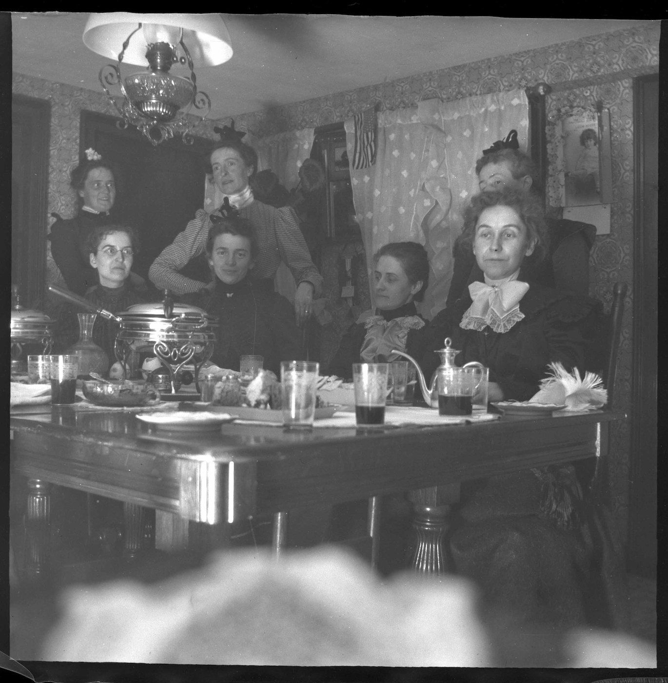 A black and white photograph of a group of people seated around a long dining table in a room with patterned wallpaper and a chandelier.