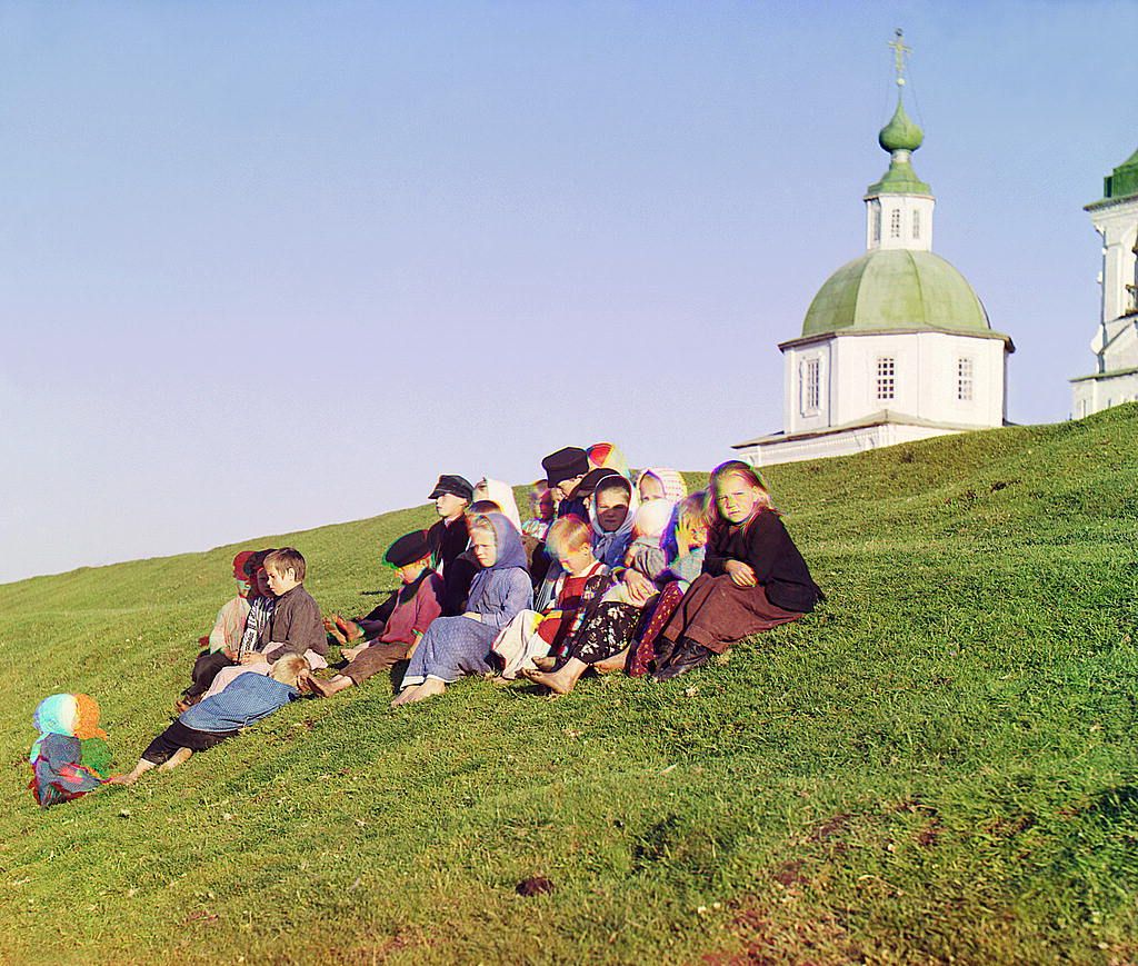 A group of children sitting on a grassy hill in front of a small church.