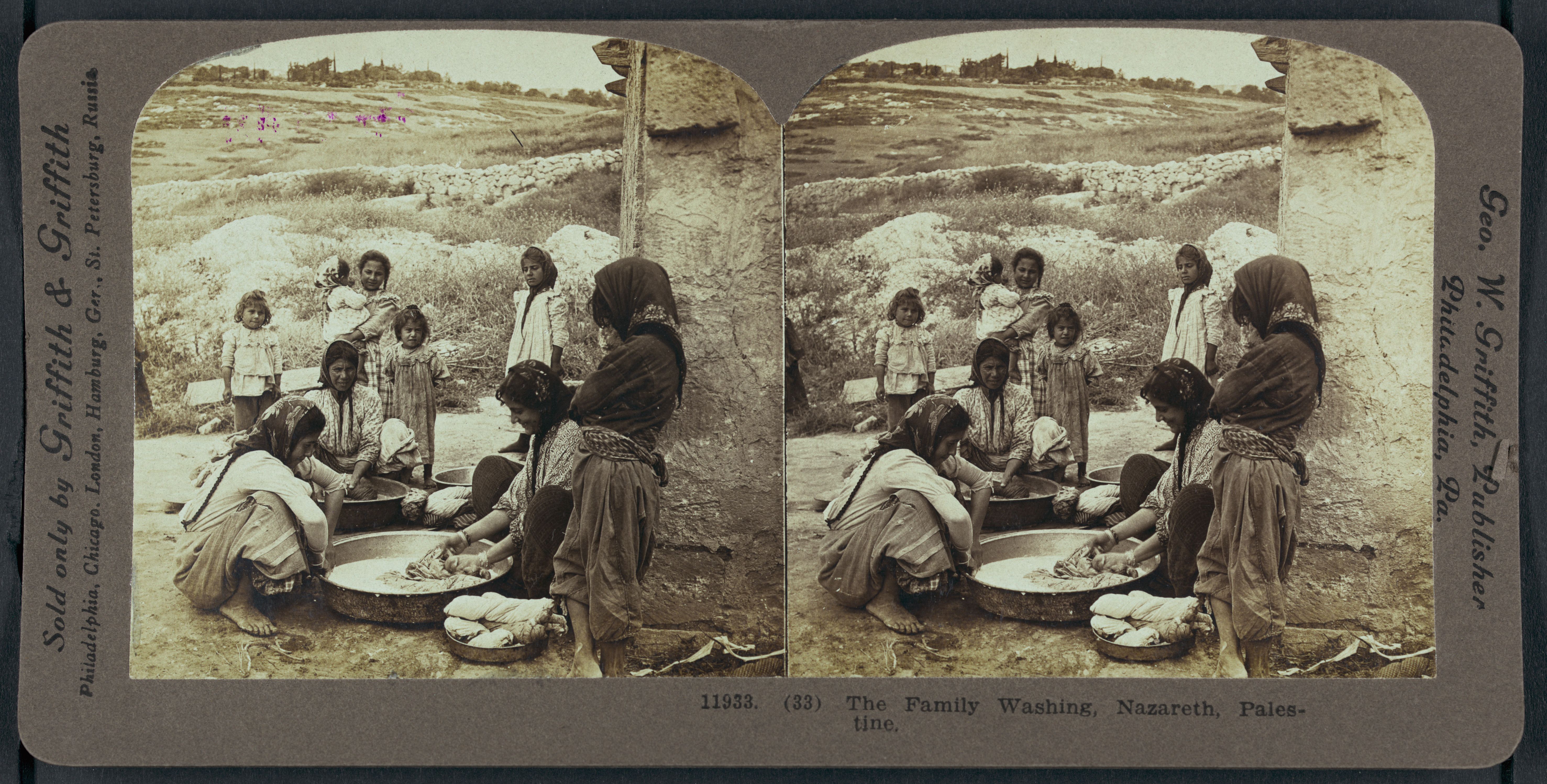 Stereoscope card depicting a family washing clothes in Nazareth, Palestine. A group of people, mostly women and children, are gathered around large basins, scrubbing, rinsing, and wringing out clothes in a rocky, arid landscape.