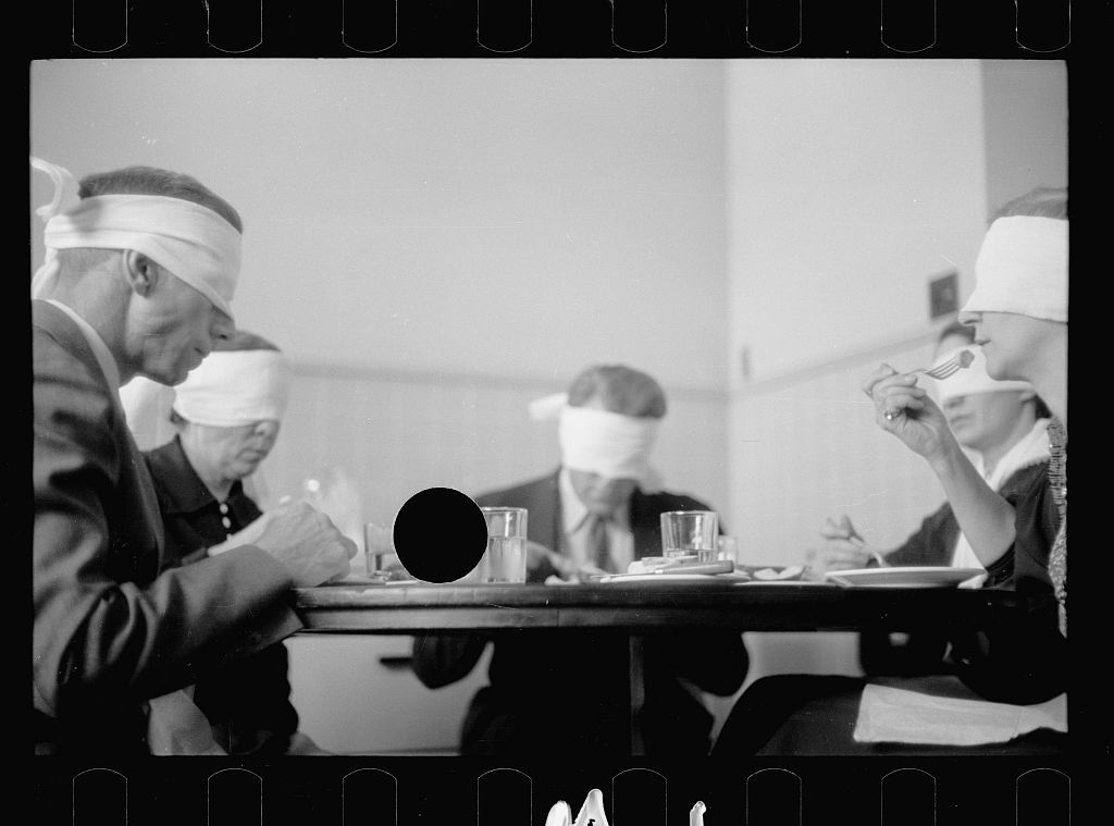 A black and white photograph of three people wearing blindfolds and paper hats, seated around a table and eating or drinking.