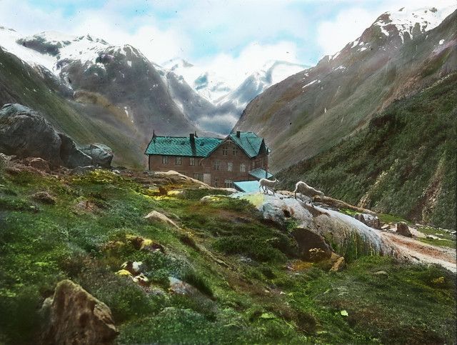 A picturesque alpine landscape featuring a wooden building with a green roof nestled between towering mountains covered in vegetation and snow.