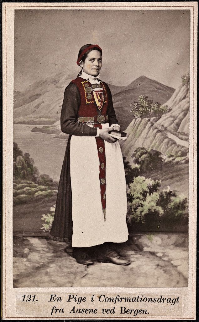 Vintage photograph of a young woman in traditional Norwegian confirmation attire, standing in a studio with a painted mountainous landscape background.