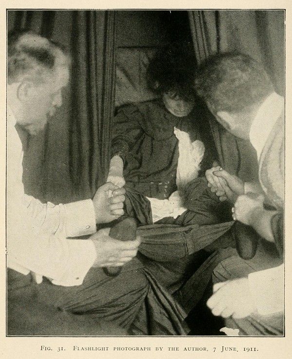 A grainy, black and white photograph taken in a dimly lit room, depicting a scene around a bed with several people gathered around a central figure who is lying down.