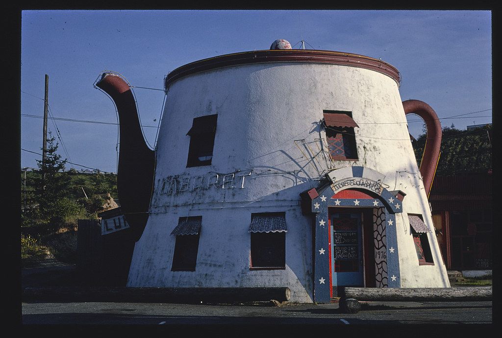 A whimsical building shaped like a giant teapot stands on a hill, with a patriotic-themed door and several windows. The sky is pale blue, and the surrounding landscape includes grassy hills, utility poles, and signage.
