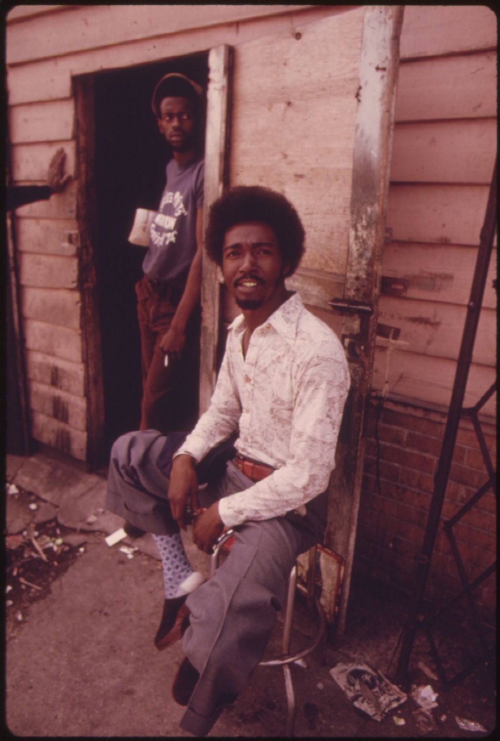 Two African American men in front of a weathered wooden building. The man in the foreground is seated on a metal chair, looking directly at the camera with a slight smile. The man in the background stands in the doorway.