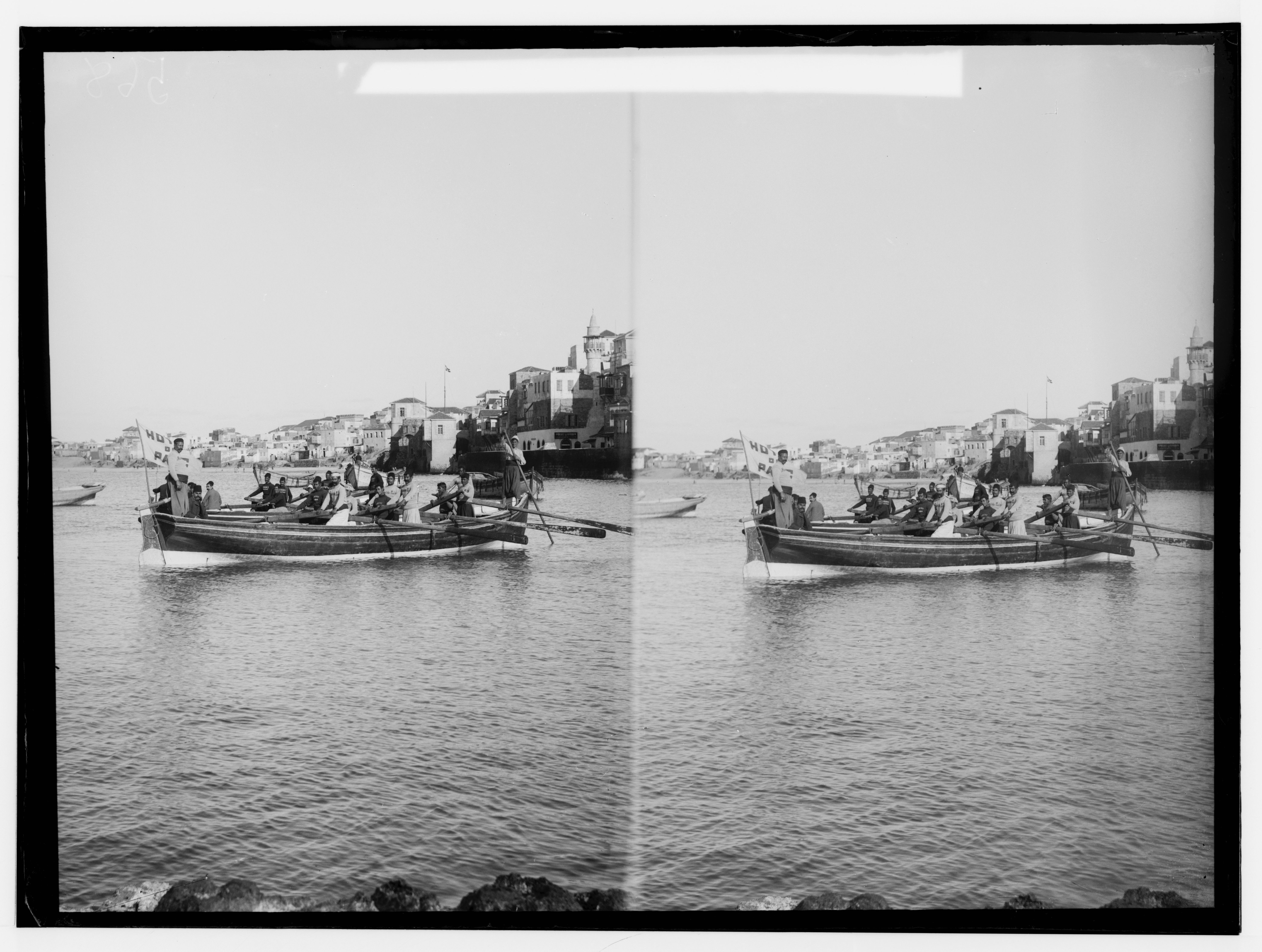 Stereoscopic photograph of two boats filled with people rowing in front of a coastal town.