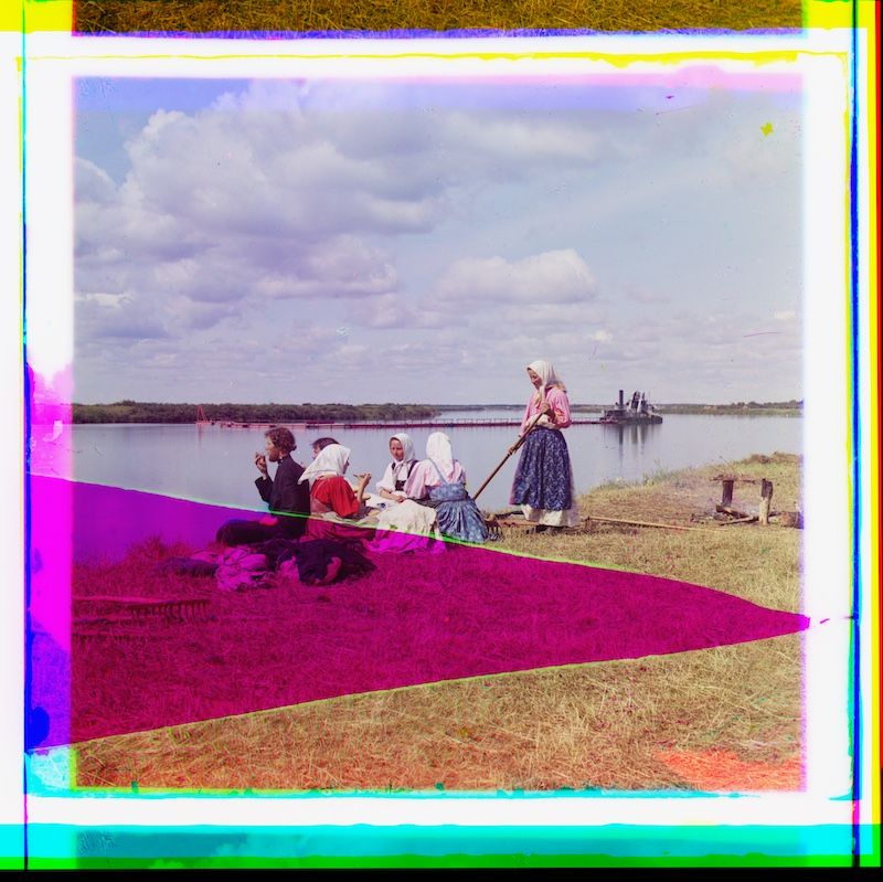 A vintage photograph of a group of people working with grain near a river or lake. The foreground is dominated by a large expanse of purple-red grain, with people seated and working on it. The background features water, boats, and a cloudy sky.