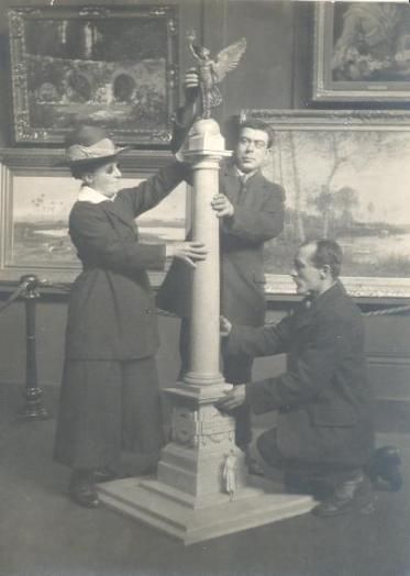 Vintage black and white photograph of three people working on a tall, ornate column in an art studio.