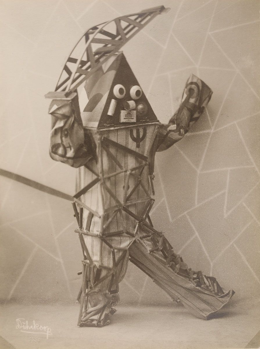 A black and white photograph of a person wearing a geometric costume made of triangles and lines, with a pyramid-shaped headpiece and stylized limbs.