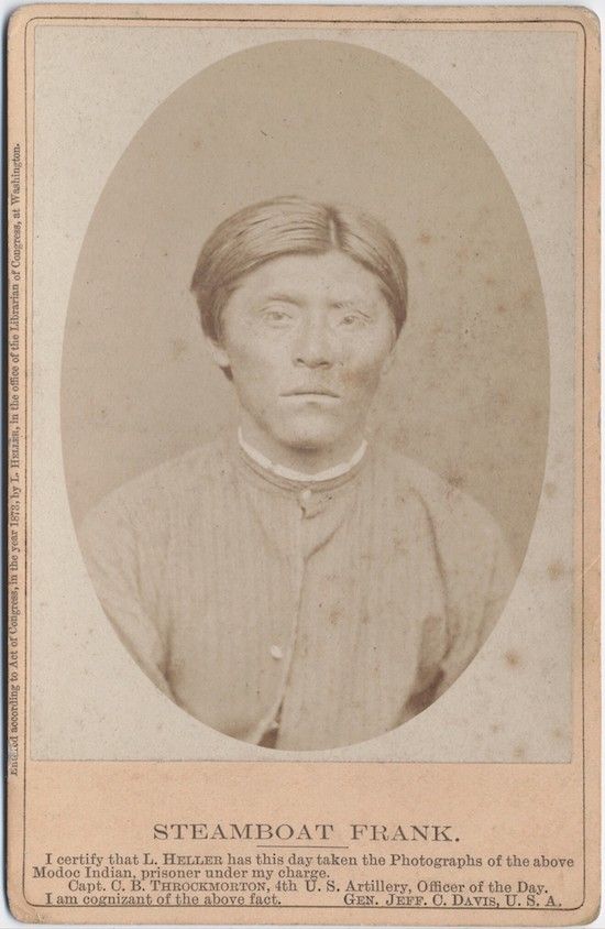 A vintage photograph of a man named Steamboat Frank, identified as a Modoc prisoner, wearing a dark jacket and light shirt. The photograph is mounted on a card with text certifying its authenticity and the photographer's information.